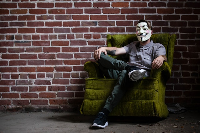 Person wearing a mask while seated against a brick wall, looking away in silence.