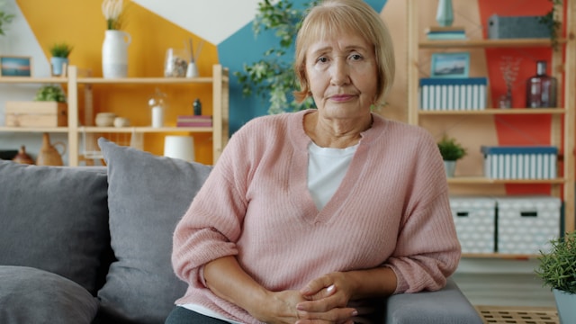 Older woman in a pink sweater sitting in her living room with a serious, reflective expression.