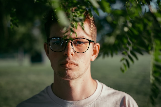 Young man with glasses standing partly behind tree branches, face partly obscured.