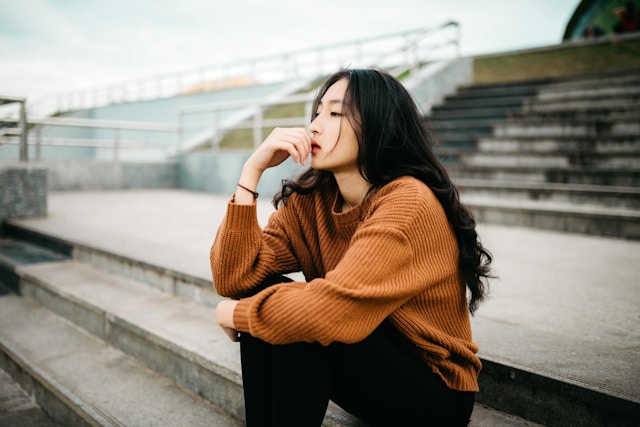 Woman in orange sweater sitting on outdoor steps looking worried and contemplative