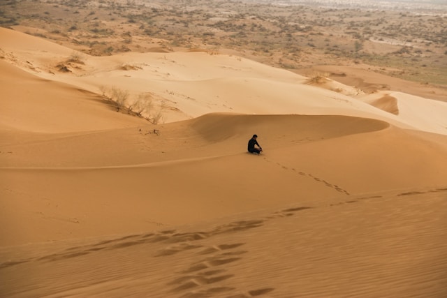A lone person sitting in the middle of wide desert sand dunes.