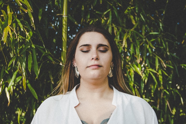 Woman standing outdoors in front of bamboo with eyes closed, practicing mindfulness.
