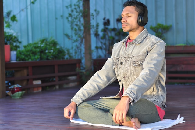 Man meditating outdoors on a deck, wearing headphones, sitting cross-legged with eyes closed.