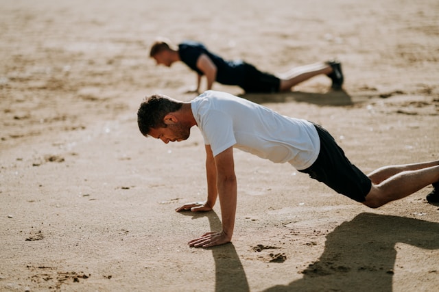 Two men doing push-ups on the beach in the sand.