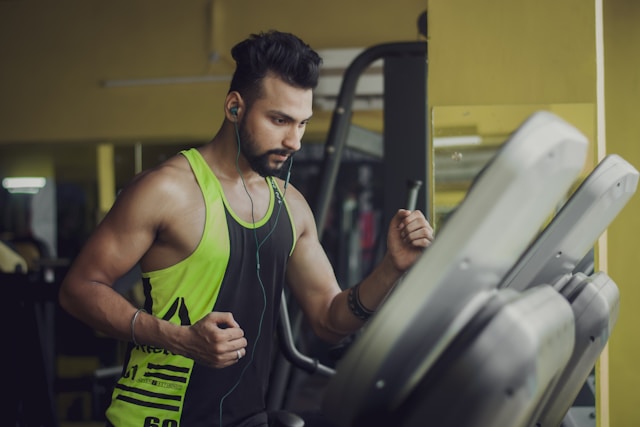 Man working out on a treadmill in a gym, wearing earbuds and tank top.
