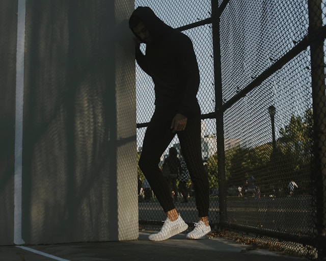 Man in a hoodie leaning against a wall by a fence, head down in distress.
