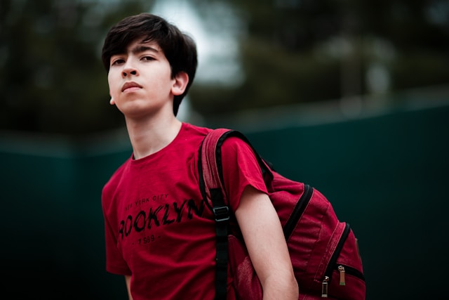 eenage boy wearing red Brooklyn t-shirt and backpack looking upward outdoors